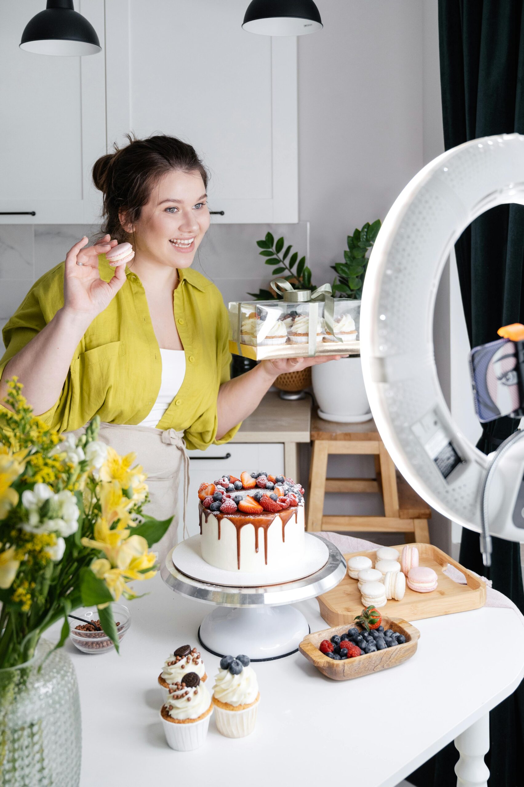 Cheerful young female confectioner smiling and demonstrating appetizing macaroons and box with cakes while filming vlog on smartphone in light kitchen